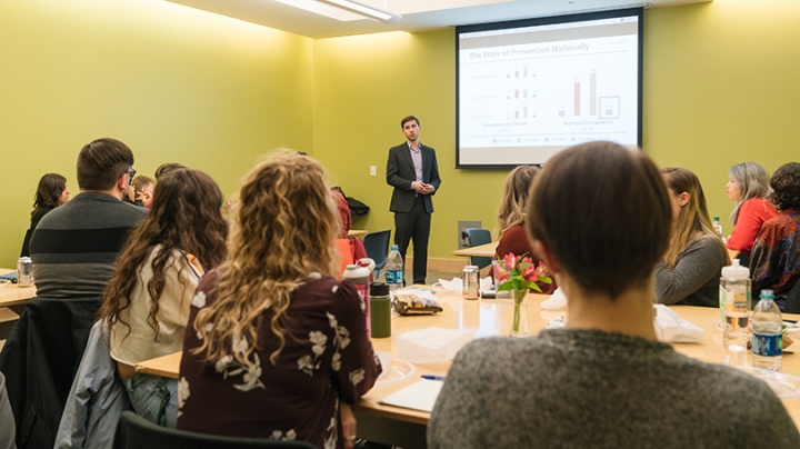 a man in a suit giving a presentation to a group of people seated in a classroom